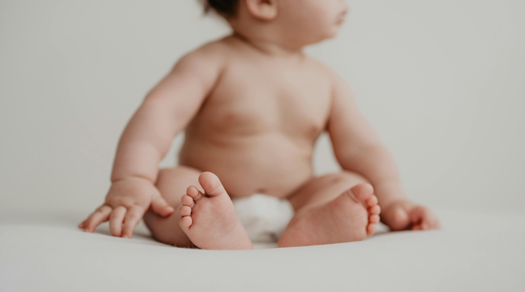 Minimalist editorial lifestyle photography of a seated baby on a white background, highlighting soft skin and natural infant wellness.