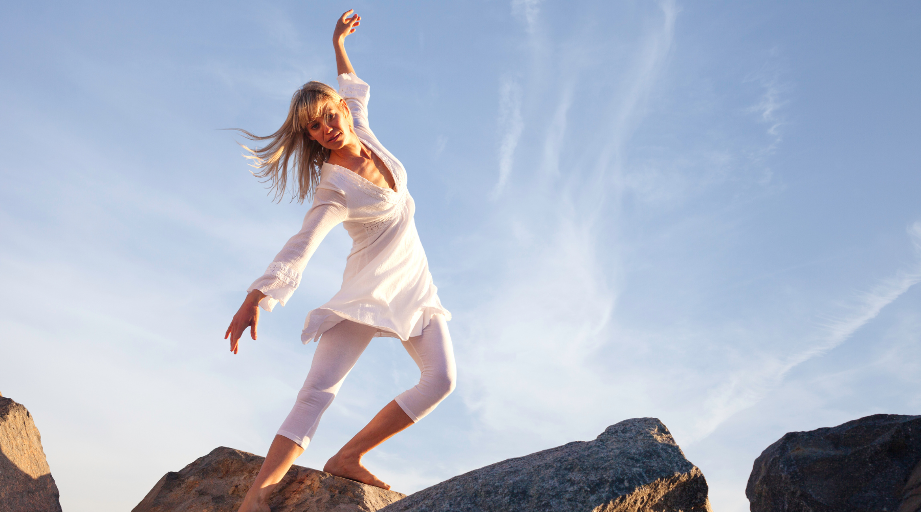 Woman in airy white outfit practicing intuitive dance on ocean rocks, representing spiritual wellness and freedom of movement.