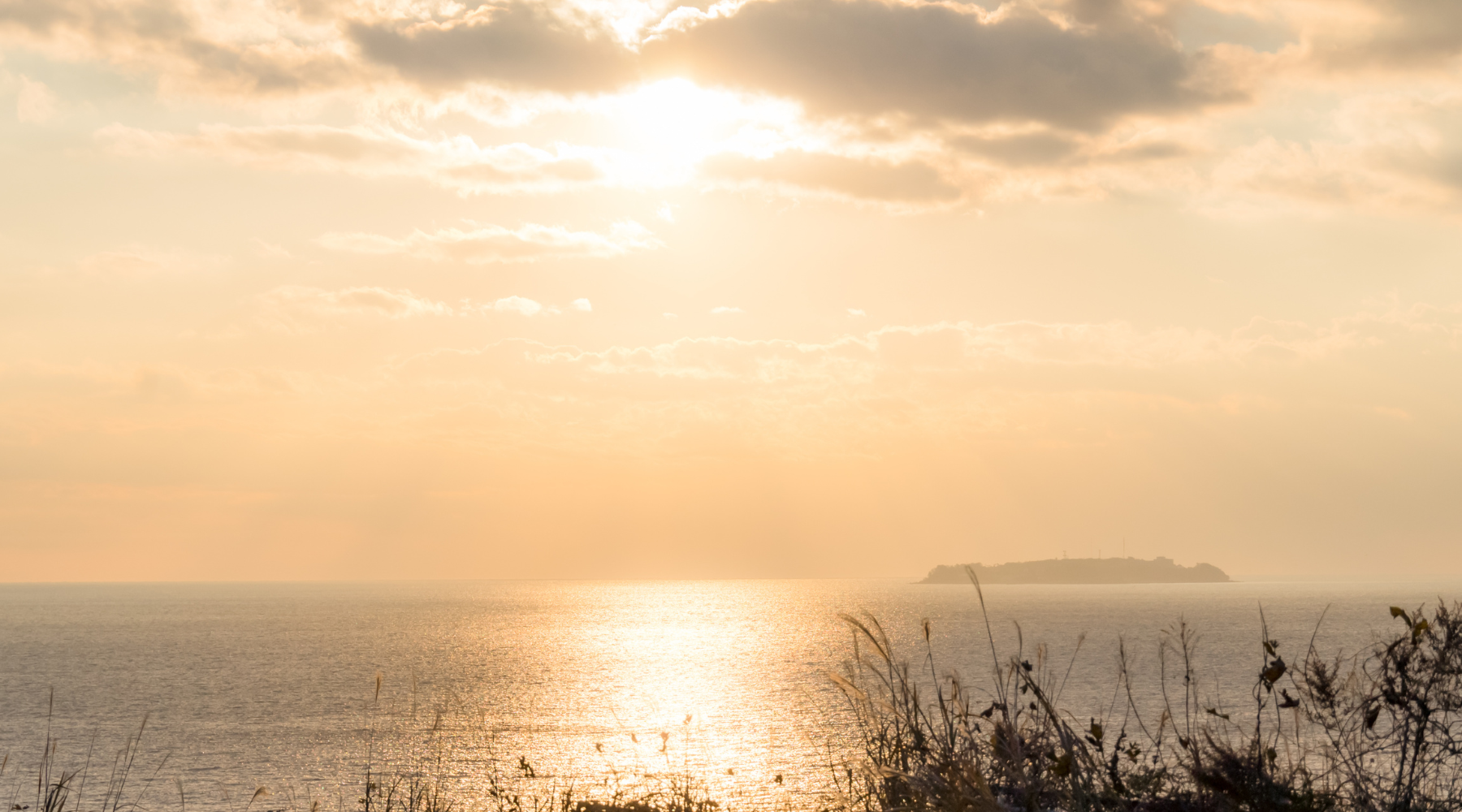 A serene, high-end photograph of a vast ocean horizon at golden hour. The sun is a soft, glowing orb diffused by light clouds, casting a shimmering path of warm light across the calm water.